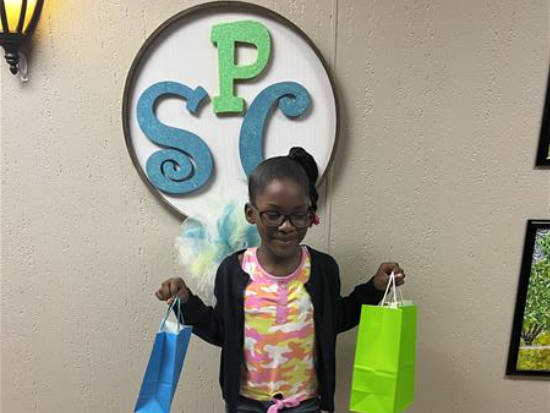 Little girl standing in front of the SPC Sign, holding up two gift bags